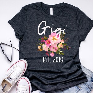 May include: A dark gray t-shirt with the text "Gigi Est. 2018" and a floral design in pink, yellow, and green.