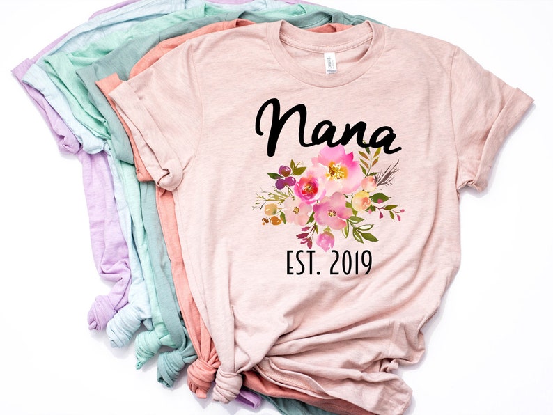 Puede incluir: Una camiseta rosa con un dise&ntilde;o floral y el texto "Nana Est. 2019" impreso en ella.