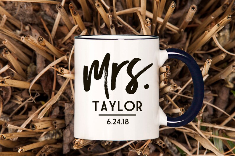 May include: White ceramic mug with a black rim and a navy blue handle. The mug has black text that says "Mrs. TAYLOR 6.24.18".