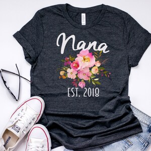Puede incluir: Una camiseta gris oscura con un dise&ntilde;o floral blanco y el texto "Nana Est. 2018".