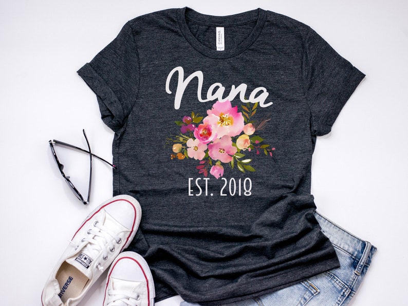 Puede incluir: Una camiseta gris oscura con un dise&ntilde;o floral blanco y el texto "Nana Est. 2018".