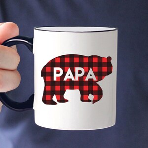 Puede incluir: Taza de cer&aacute;mica blanca con asa y borde negros. La taza presenta una silueta de oso en patr&oacute;n de cuadros rojos y negros con la palabra "PAPA" en letras blancas.