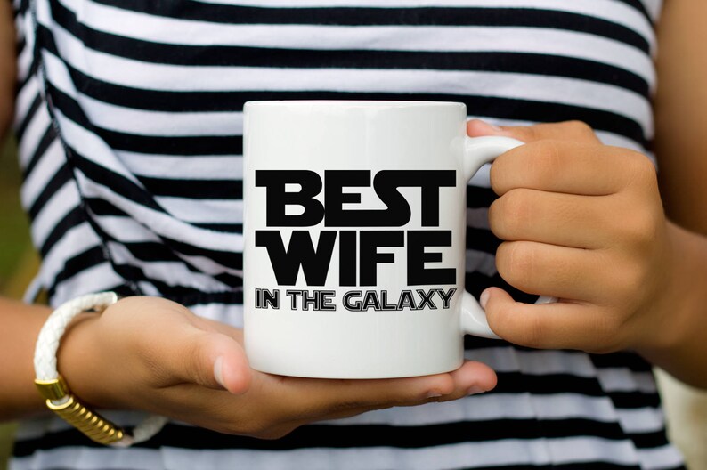 May include: White ceramic mug with the text "BEST WIFE IN THE GALAXY" in black block letters. The mug is held by a person wearing a black and white striped top and a white bracelet.