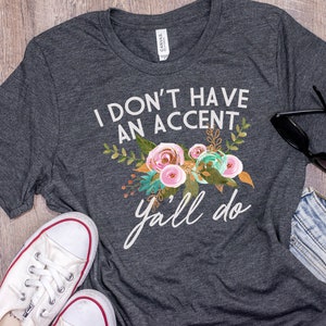 以下が含まれることがあります： 白い花の模様と「I don't have an accent y'all do」という文字が入った濃い灰色のTシャツです。