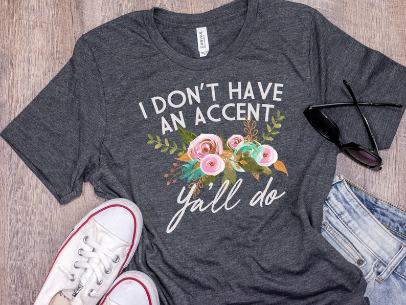 以下が含まれることがあります： 白い花の模様と「I don't have an accent y'all do」という文字が入った濃い灰色のTシャツです。