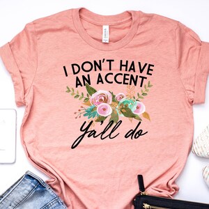 以下が含まれることがあります： ピンクのTシャツに「I don't have an accent y'all do」という黒い文字が書かれています。テキストは、ピンク、グリーン、ゴールドのアクセントがついた花柄のデザインで囲まれています。