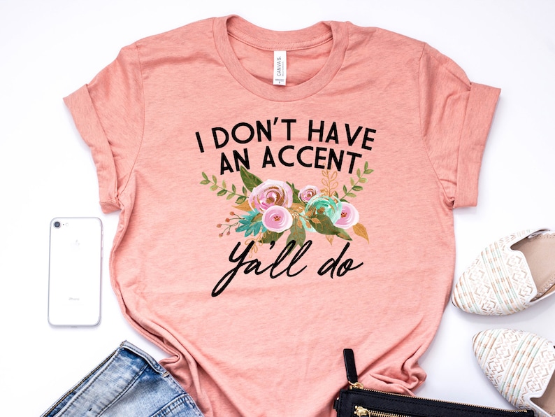 以下が含まれることがあります： ピンクのTシャツに「I don't have an accent y'all do」という黒い文字が書かれています。テキストは、ピンク、グリーン、ゴールドのアクセントがついた花柄のデザインで囲まれています。
