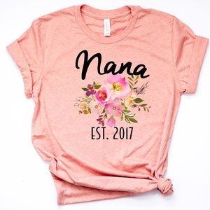 Puede incluir: Una camiseta de color melocot&oacute;n con la palabra "Nana" en letras cursivas negras y un dise&ntilde;o floral en rosa, amarillo y morado. El texto "EST. 2017" est&aacute; impreso debajo del dise&ntilde;o floral.