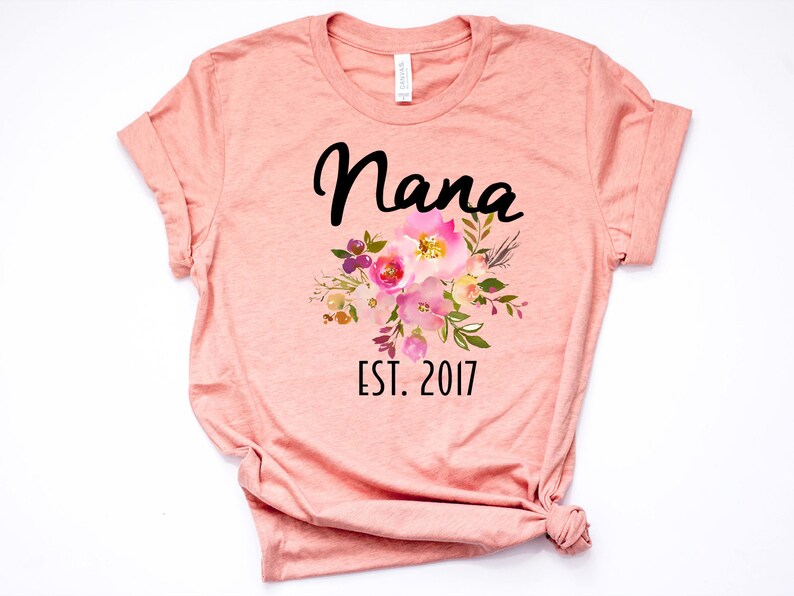 Puede incluir: Una camiseta de color melocot&oacute;n con la palabra "Nana" en letras cursivas negras y un dise&ntilde;o floral en rosa, amarillo y morado. El texto "EST. 2017" est&aacute; impreso debajo del dise&ntilde;o floral.