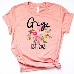 May include: A peach-colored t-shirt with the text "Gigi Est. 2021" and a floral design in pink, yellow, and green.