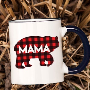 Puede incluir: Taza de cer&aacute;mica blanca con borde y asa azules. La taza presenta una silueta de oso de cuadros rojos y negros con la palabra "MAMA" impresa dentro del oso.