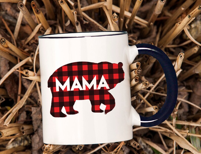 Puede incluir: Taza de cer&aacute;mica blanca con borde y asa azules. La taza presenta una silueta de oso de cuadros rojos y negros con la palabra "MAMA" impresa dentro del oso.