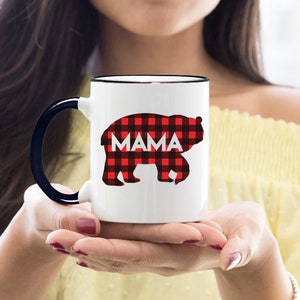 Puede incluir: Taza de cer&aacute;mica blanca con borde y asa negros. La taza presenta una silueta de oso de cuadros rojos y negros con la palabra "MAMA" impresa en blanco.