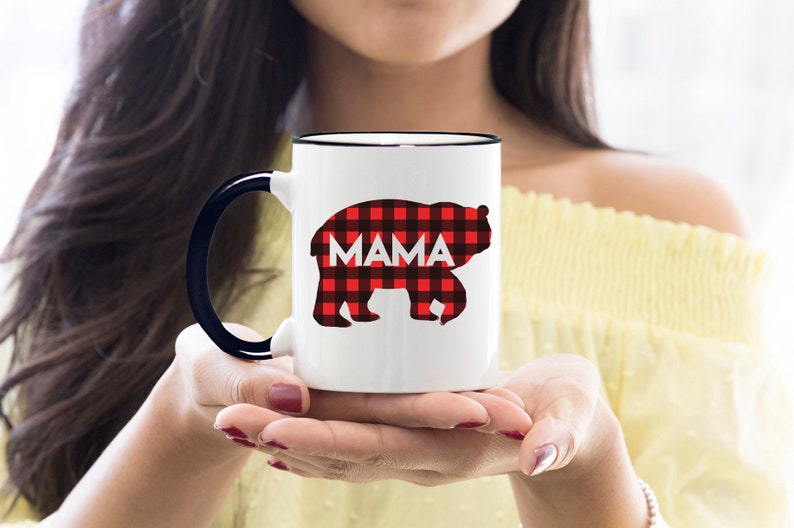 Puede incluir: Taza de cer&aacute;mica blanca con borde y asa negros. La taza presenta una silueta de oso de cuadros rojos y negros con la palabra "MAMA" impresa en blanco.