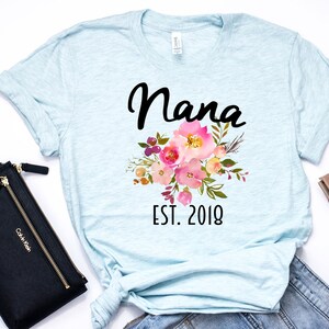 Puede incluir: Camiseta azul claro con un dise&ntilde;o floral y el texto "Nana Est. 2018" impreso en la parte delantera.