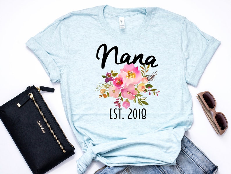 Puede incluir: Camiseta azul claro con un dise&ntilde;o floral y el texto "Nana Est. 2018" impreso en la parte delantera.