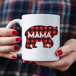 Puede incluir: Taza de cer&aacute;mica blanca con una silueta de oso de cuadros rojos y negros. La palabra "MAMA" est&aacute; impresa en blanco dentro de la silueta del oso.