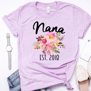 Puede incluir: Una camiseta lavanda con un dise&ntilde;o floral y el texto "Nana Est. 2018".