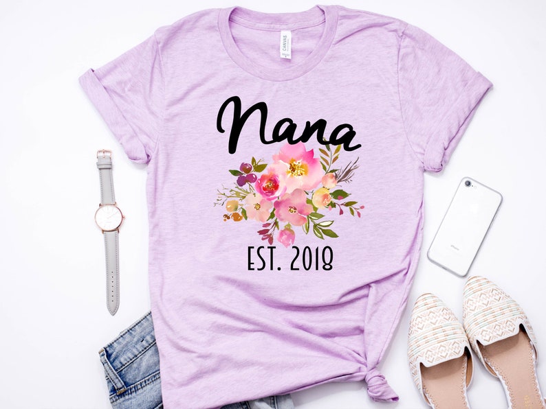 Puede incluir: Una camiseta lavanda con un dise&ntilde;o floral y el texto "Nana Est. 2018".