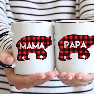 Puede incluir: Dos tazas de cer&aacute;mica blanca con asas negras. Las tazas tienen un dise&ntilde;o de cuadros de b&uacute;falo rojo y negro con las palabras "Mama" y "Papa" en ellas.