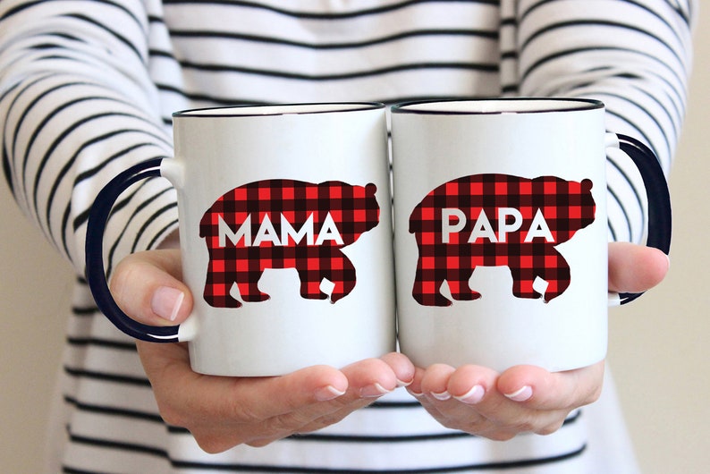 Puede incluir: Dos tazas de cer&aacute;mica blanca con asas negras. Las tazas tienen un dise&ntilde;o de cuadros de b&uacute;falo rojo y negro con las palabras "Mama" y "Papa" en ellas.