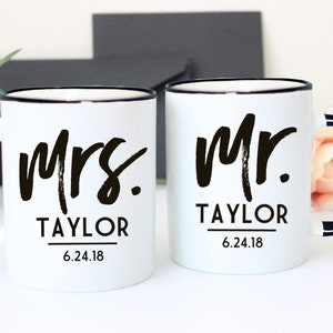 May include: Two white ceramic mugs with black rims and handles. The mug on the left says "Mrs. Taylor 6.24.18" and the mug on the right says "Mr. Taylor 6.24.18".