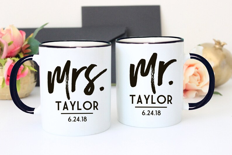 May include: Two white ceramic mugs with black rims and handles. The mug on the left says "Mrs. Taylor 6.24.18" and the mug on the right says "Mr. Taylor 6.24.18".
