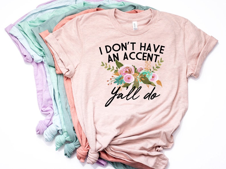 以下が含まれることがあります： 「I don't have an accent y'all do」というテキストと花柄のデザインが施されたピンクのTシャツです。