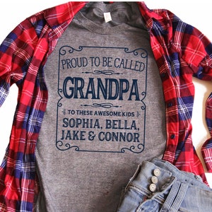 May include: Gray t-shirt with a vintage-style design that says "Proud to be called Grandpa" with the names Sophia, Bella, Jake & Connor underneath. The shirt is layered over a red and blue plaid shirt.