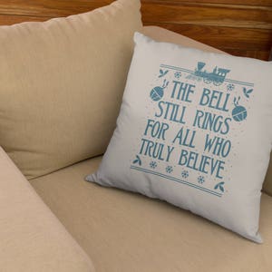 May include: A white throw pillow with blue text that reads "The Bell Still Rings For All Who Truly Believe". The text is surrounded by a blue snowflake pattern. A blue train with a bell is pictured above the text.