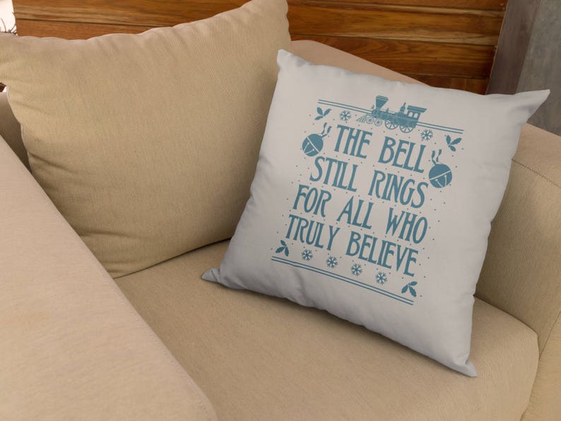 May include: A white throw pillow with blue text that reads "The Bell Still Rings For All Who Truly Believe". The text is surrounded by a blue snowflake pattern. A blue train with a bell is pictured above the text.