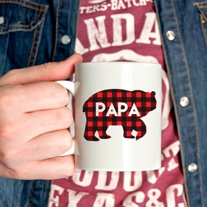 Puede incluir: Taza de cer&aacute;mica blanca con una silueta de oso de cuadros rojos y negros. El oso tiene la palabra "PAPA" escrita en blanco en su pecho.