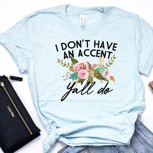 以下が含まれることがあります： 薄い青色のTシャツに花柄のデザインと「I don't have an accent y'all do」というテキストが書かれています。