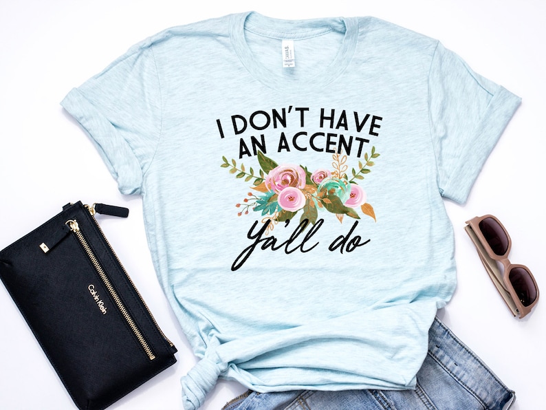 以下が含まれることがあります： 薄い青色のTシャツに花柄のデザインと「I don't have an accent y'all do」というテキストが書かれています。