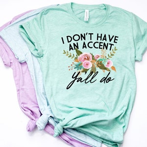 以下が含まれることがあります： 花柄のライトグリーンのTシャツに、黒い文字で「I don't have an accent y'all do」と書いてあります。