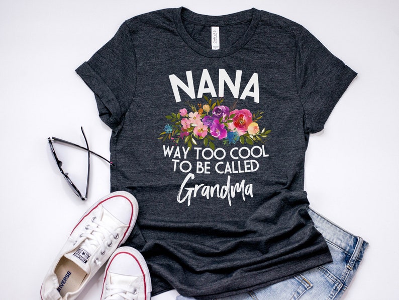 Puede incluir: Una camiseta gris oscuro con un dise&ntilde;o floral blanco y el texto "NANA WAY TOO COOL TO BE CALLED GRANDMA".