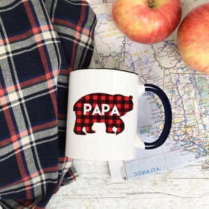 Puede incluir: Taza de cer&aacute;mica blanca con borde y asa negros. La taza tiene un oso de cuadros rojos y negros con la palabra "PAPA" impresa.