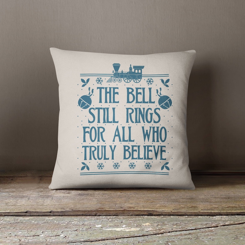 May include: A white throw pillow with blue text that reads "The Bell Still Rings For All Who Truly Believe". The text is surrounded by a blue train and snowflakes.