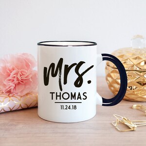 May include: White ceramic mug with a black rim and handle. The mug has black text that says "Mrs. THOMAS 11.24.18".