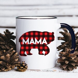 Puede incluir: Taza de cer&aacute;mica blanca con borde y asa negros. La taza presenta una silueta de oso de cuadros rojos y negros con la palabra "MAMA" impresa en blanco.