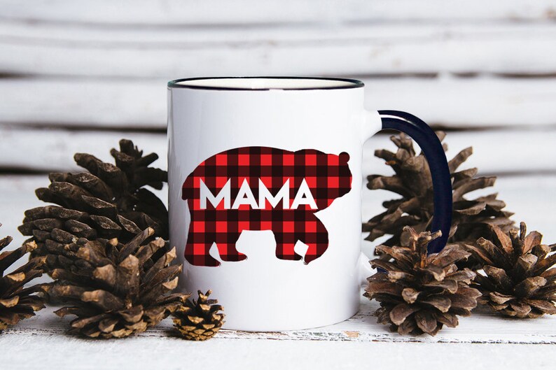 Puede incluir: Taza de cer&aacute;mica blanca con borde y asa negros. La taza presenta una silueta de oso de cuadros rojos y negros con la palabra "MAMA" impresa en blanco.