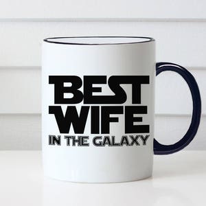 May include: White ceramic mug with a black rim and handle. The mug features the text "BEST WIFE IN THE GALAXY" in bold black letters, a fun and playful design. The mug is a great gift idea.