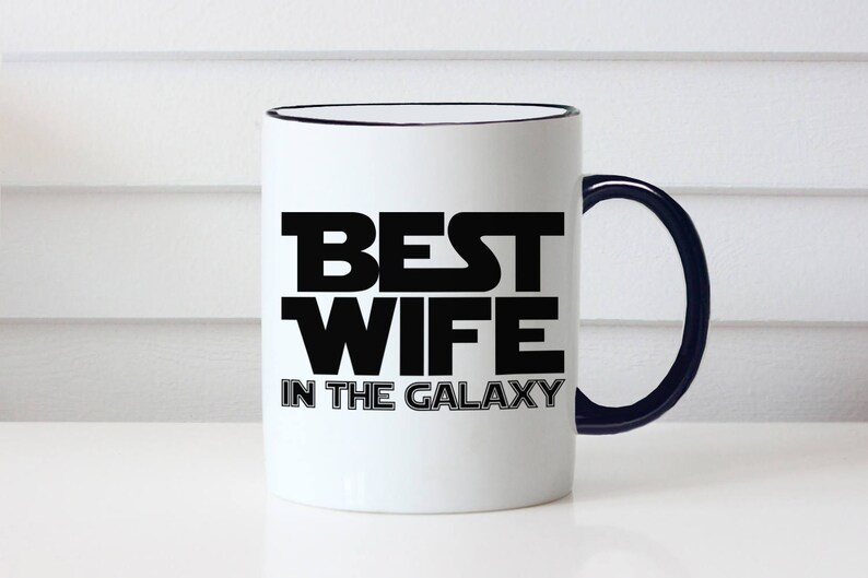 May include: White ceramic mug with a black rim and handle. The mug features the text "BEST WIFE IN THE GALAXY" in bold black letters, a fun and playful design. The mug is a great gift idea.
