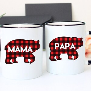 Puede incluir: Dos tazas de cer&aacute;mica blanca con bordes y asas negros. Las tazas presentan una silueta de oso de cuadros rojos y negros con las palabras "Mama" y "Papa" impresas en ellas.