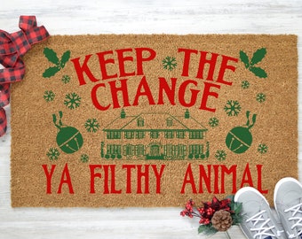 以下が含まれることがあります： 赤い文字で「KEEP THE CHANGE YA FILTHY ANIMAL」と書かれた茶色のココナッツ繊維のドアマット。ドアマットには緑色の家のイラストと緑色の鈴が2つ描かれています。