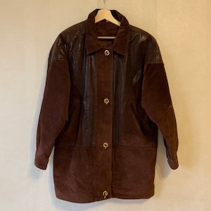 May include: A brown suede jacket with leather accents and gold buttons. The jacket has a collar and long sleeves.