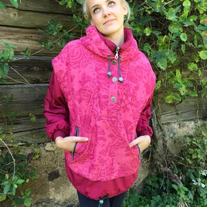 May include: A pink and red patterned jacket with a high collar and a snap closure. The jacket has a drawstring hood and two pockets.
