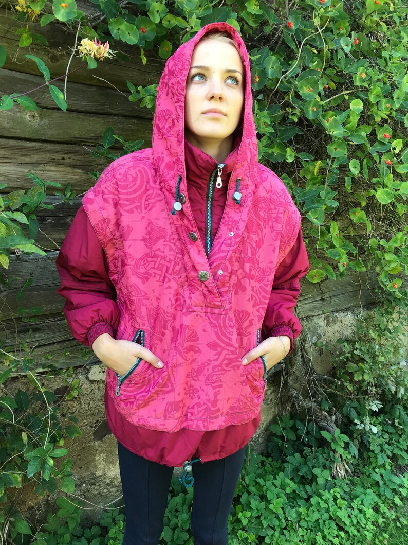 May include: A pink hooded jacket with a patterned design and a zipper closure. The jacket has two pockets on the front and a drawstring at the bottom.