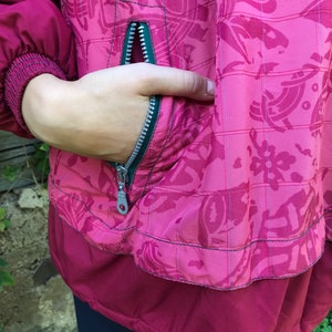 May include: A burgundy jacket with a green zipper and a floral pattern. The jacket has a pocket on the front with a zipper.