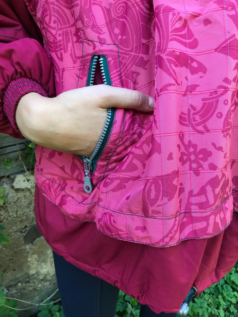 May include: A burgundy jacket with a green zipper and a floral pattern. The jacket has a pocket on the front with a zipper.